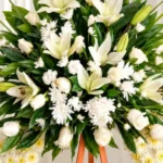 Detalle del copete de la Corona Fúnebre Espinas de Consuelo: lirios, rosas y crisantemos blancos entre follaje verde, sobre la cinta de condolencia en letras doradas.
