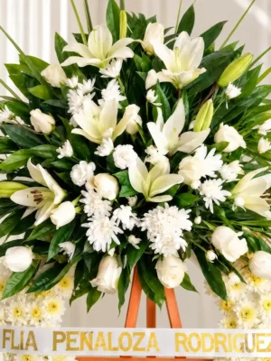Detalle del copete de la Corona Fúnebre Espinas de Consuelo: lirios, rosas y crisantemos blancos entre follaje verde, sobre la cinta de condolencia en letras doradas.