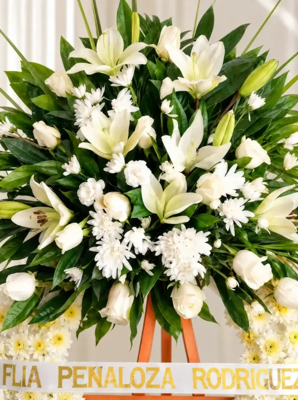 Detalle del copete de la Corona Fúnebre Espinas de Consuelo: lirios, rosas y crisantemos blancos entre follaje verde, sobre la cinta de condolencia en letras doradas.