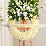Corona Fúnebre Espinas de Consuelo con base de margaritas blancas y copete de lirios, rosas y follaje, sobre atril de madera.
