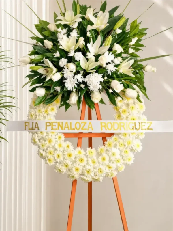 Corona Fúnebre Espinas de Consuelo con base de margaritas blancas y copete de lirios, rosas y follaje, sobre atril de madera.