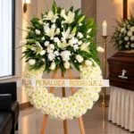 Corona Fúnebre Espinas de Consuelo sobre atril de madera en una sala de velación: base de margaritas blancas con copete de lirios, rosas y crisantemos blancos y cinta de condolencia, junto al cofre y una cruz.