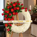 Corona Fúnebre Eternidad sobre atril de madera en una sala de velación: crisantemos blancos con una cascada de rosas y gerberas rojas y cinta de condolencia, junto al cofre y una cruz.