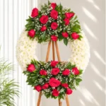 Corona Fúnebre Lazos de Amor circular de crisantemos blancos con rosas rojas y fucsia arriba y abajo, sobre atril de madera.