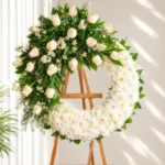 Corona Fúnebre Luz Eterna circular de crisantemos blancos con rosas crema y margaritas arriba, sobre atril de madera.