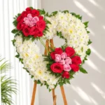 Corona Fúnebre Ternura en forma de corazón de crisantemos blancos con rosas rojas y rosadas, sobre atril de madera.