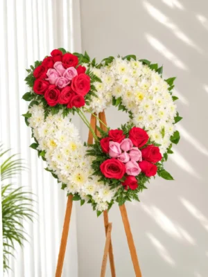 Corona Fúnebre Ternura en forma de corazón de crisantemos blancos con rosas rojas y rosadas, sobre atril de madera.
