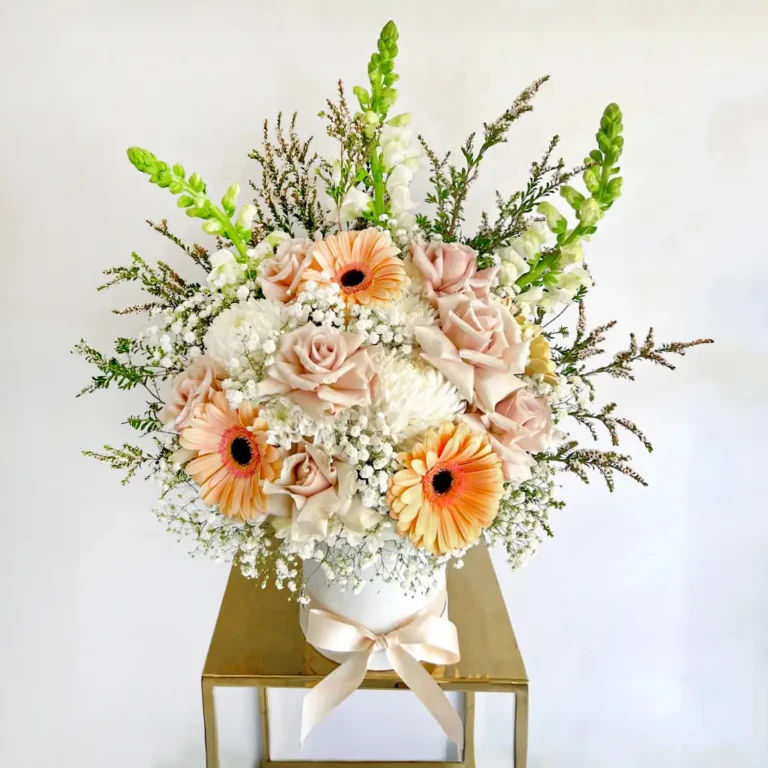 Ramo de flores con gerberas melocotón, rosas crema y follaje, en base blanca con lazo, ideal para regalar con elegancia.