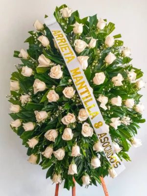 Pedestal fúnebre ovalado con rosas color crema y follaje verde, con cinta de dedicatoria sobre atril de madera