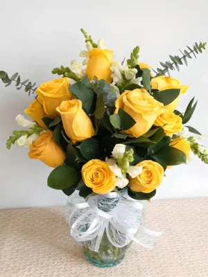 Ramo fúnebre en florero de vidrio con rosas amarillas, flores blancas pequeñas y follaje de eucalipto