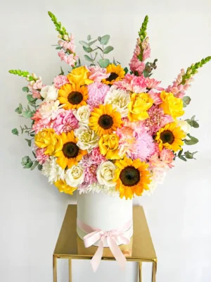 Ramo de girasoles con rosas blancas y amarillas, flores rosadas y follaje en base cilíndrica con lazo rosado.