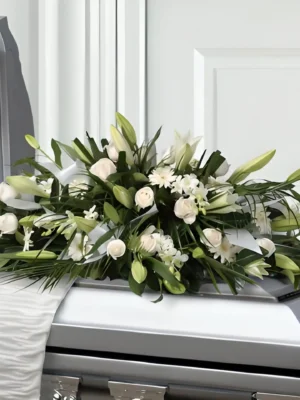 Cubre cofre alargado con lirios y rosas blancas entre follaje verde, sobre un ataúd plateado