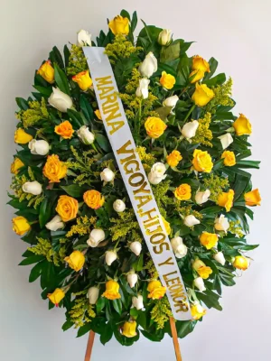 Pedestal fúnebre ovalado con rosas amarillas, naranjas y blancas entre follaje verde, sobre atril de madera