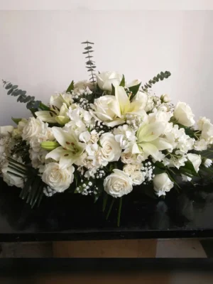 Cubre cofre fúnebre ovalado con rosas y lirios blancos entre ramas de eucalipto, sobre un cofre oscuro
