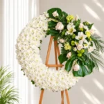 Corona Fúnebre Ágata circular de crisantemos blancos con rosas crema, lirios blancos y follaje verde, sobre atril de madera.