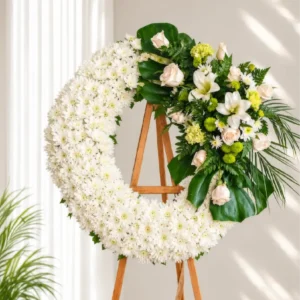 Corona Fúnebre Ágata circular de crisantemos blancos con rosas crema, lirios blancos y follaje verde, sobre atril de madera.
