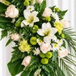 Detalle de la Corona Fúnebre Ágata: rosas crema, lirios blancos, claveles y pompones verde limón y margaritas blancas entre helecho y hojas de palma.