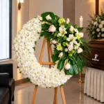 Corona Fúnebre Ágata sobre atril de madera en una sala de velación: crisantemos blancos con un ramillete lateral de rosas crema, lirios blancos y flores verdes, junto al cofre y una cruz.