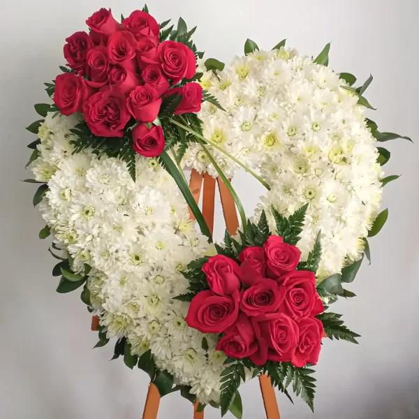 Corona Fúnebre Armonía Eterna en forma de corazón de crisantemos blancos con dos ramilletes de rosas rojas y helecho, sobre atril de madera.