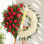 Corona Fúnebre Aurora circular, mitad rosas rojas con helecho y mitad crisantemos blancos, con cinta blanca de dedicatoria, sobre atril de madera.