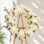 Corona Fúnebre Celeste circular de crisantemos y margaritas blancas con rosas crema, un lirio y eucalipto, sobre atril de madera.