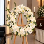 Corona Fúnebre Celeste sobre atril de madera en una sala de velación: corona circular de rosas crema, lirios, margaritas y crisantemos blancos con eucalipto, junto al cofre y una cruz.