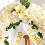 Detalle de la Corona Fúnebre Cristal: lirios blancos, crisantemos blancos de centro amarillo, rosas crema y durazno, claveles blancos y helecho, con cinta blanca de condolencia en letras doradas.