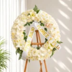 Corona Fúnebre Cristal circular de rosas, claveles y lirios blancos, con cinta de condolencia, sobre atril de madera.
