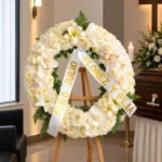 Corona Fúnebre Cristal sobre atril de madera en una sala de velación: crisantemos blancos con lirios, rosas crema y durazno y claveles blancos, cinta de condolencia en letras doradas, junto al cofre y una cruz.