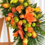Detalle de la Corona Fúnebre Descansa en Paz: rosas y lirios naranjas, pompones verdes y solidago amarillo entre hojas de palma.
