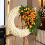 Corona Fúnebre Descansa en Paz sobre atril de madera en una sala de velación: crisantemos blancos en media luna con un ramillete de rosas y lirios naranjas y pompones verdes, junto al cofre y una cruz.