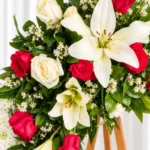 Detalle de la Corona Fúnebre Doncella: lirios blancos, rosas rojas y blancas y pequeñas flores blancas entre follaje verde.