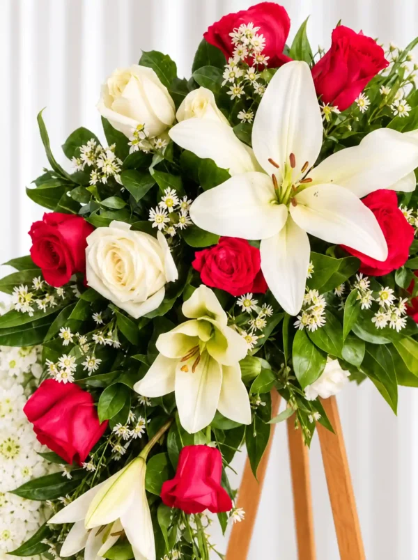 Detalle de la Corona Fúnebre Doncella: lirios blancos, rosas rojas y blancas y pequeñas flores blancas entre follaje verde.