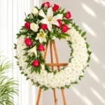Corona Fúnebre Doncella de crisantemos blancos con ramillete de lirios blancos, rosas rojas y blancas, sobre atril de madera.