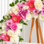 Detalle de la Corona Fúnebre Elegía de Colores: gerberas coral, rosas lila y blancas, margaritas rosadas y crisantemos blancos, con la cinta de condolencia en letras doradas.