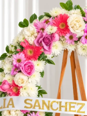 Detalle de la Corona Fúnebre Elegía de Colores: gerberas coral, rosas lila y blancas, margaritas rosadas y crisantemos blancos, con la cinta de condolencia en letras doradas.