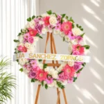 Corona Fúnebre Elegía de Colores, con crisantemos blancos, gerberas coral y rosadas, y rosas lila y fucsia, sobre atril de madera.