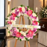 Corona Fúnebre Elegía de Colores sobre atril de madera en una sala de velación: corona circular de crisantemos blancos, gerberas coral, rosas lila y margaritas rosadas, con cinta de condolencia, junto al cofre y una cruz.
