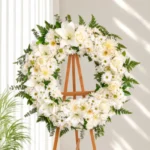 Corona Fúnebre Esmeralda de crisantemos, lirios y rosas blancas con gerberas araña y follaje verde, sobre atril de madera.