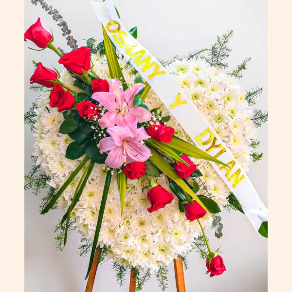 Corona Fúnebre Gema en forma de corazón de crisantemos blancos con lirios rosados, rosas rojas y gypsophila, con cinta de condolencia.