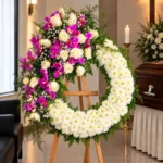 Corona Fúnebre Hechizo de Paz sobre atril de madera en una sala de velación: crisantemos blancos con una cascada de rosas blancas y orquídeas fucsia, junto al cofre y una cruz.