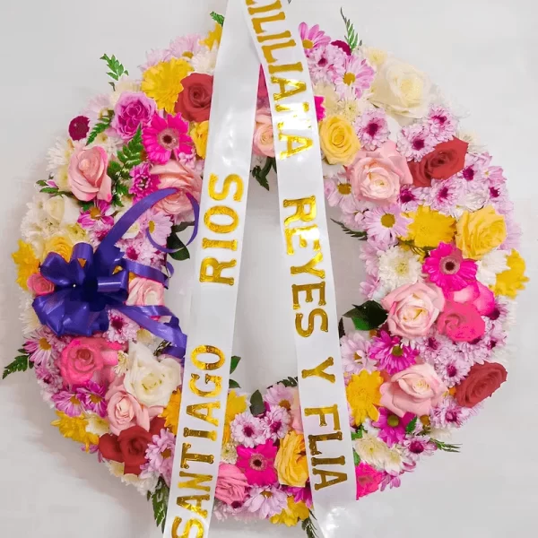 Corona Fúnebre Iris circular multicolor de rosas amarillas, rosadas y rojas, gerberas fucsia y margaritas lila, con moño morado y cintas de condolencia.