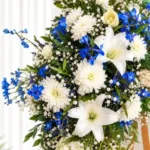 Detalle de la Corona Fúnebre Magia Eterna: lirios y crisantemos blancos, espuela de caballero azul, gypsophila y una rosa crema.