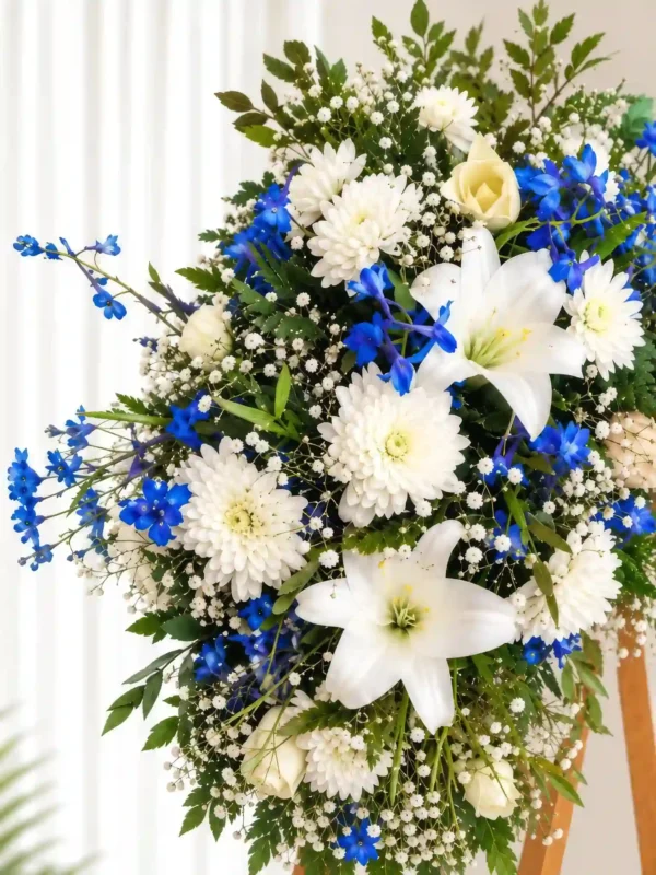 Detalle de la Corona Fúnebre Magia Eterna: lirios y crisantemos blancos, espuela de caballero azul, gypsophila y una rosa crema.