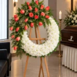 Corona Fúnebre Memoria sobre atril de madera en una sala de velación: rosas coral con caspia y crisantemos blancos, junto al cofre y una cruz.