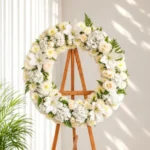 Corona Fúnebre Pureza circular de flores blancas: lirios, rosas crema, hortensias y crisantemos con helecho, sobre atril de madera.