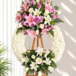 Corona Fúnebre Reencuentro circular de crisantemos blancos con lirios rosados y blancos, rosas lila y pompones fucsia arriba, y lirios y rosas blancas abajo, sobre atril de madera.