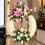 Corona Fúnebre Reencuentro sobre atril de madera en una sala de velación: crisantemos blancos con un ramillete de lirios rosados y blancos y rosas lila arriba, y lirios y rosas blancas abajo, junto al cofre y una cruz.