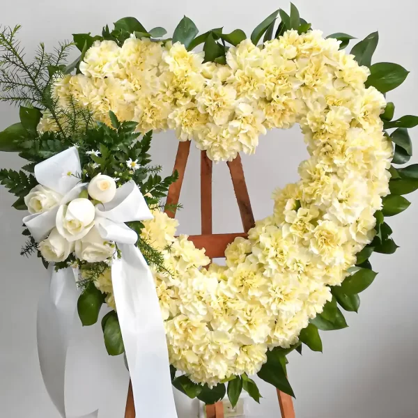 Corona Fúnebre Tranquilidad en Flores en forma de corazón, tapizada en claveles color crema, con moño blanco y rosas blancas pequeñas, sobre atril de madera.