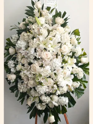 Pedestal fúnebre ovalado con rosas, lirios y astromelias blancas sobre atril de madera